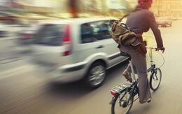 Ein Radfahrer fährt rechts auf einem Klapprad auf der Straße und wird gerade von einem weißen Auto links überholt.