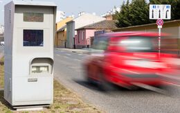 Links im Bild sieht man eine Radarbox, bei der ein rotes Auto auf der Straße vorbeifährt.