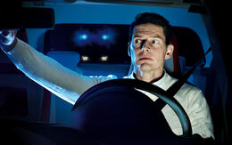 Ein junger Lenker im Auto schaut in den Rückspiegel. Hinter der Heckscheibe sieht man ein Fahrzeug mit Blaulicht.