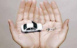 Geöffnete Hand hält in den Handflächen links ein weißes Matchbox-Auto, rechts liegt eine Person (Fotomontage) auf dem Boden.