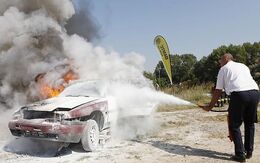 Auf einer Schotterfläche steht ein verbranntes Fahrzeug, ein Mann löscht dieses mit einem Handfeuerlöscher. Im Hintergrund sind eine ÖAMTC Fahrtechnik Fahne und Bäume.