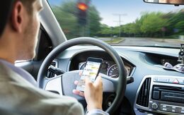 Ein Mann sitzt in einem Auto, in seiner rechten Hand hält er ein Smartphone. Sein Blick ist auf das Handy gerichtet, während das Lenkrad mit der linken Hand gehalten wird. Im Hintergrund sind das Armaturenbrett und Teile der Windschutzscheibe zu sehen. Die Szene vermittelt den Eindruck, dass die Person während der Fahrt durch das Handy abgelenkt ist.