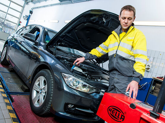 Ein dunkles Auto steht in der KFZ Werkstatt auf der flachen Hebebühne, ein KFZ Techniker steht bei der geöffneten Motorhaube und überprüft mit einem Messgerät den Motor.