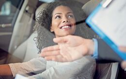 Lächelnde, junge Frau sitzt in einem Auto auf dem Fahrersitz während ein Mann mit einem Klemmbrett ihr etwas erklärt.