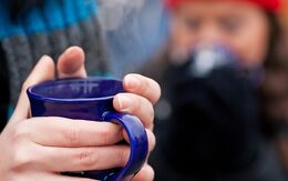 Eine Hand umfasst eine blaue Teetasse, Im Hintergrund sieht man verschwommen, dass sie einen Schal trägt bzw. eine weitere Person mit einer roten Mütze.
