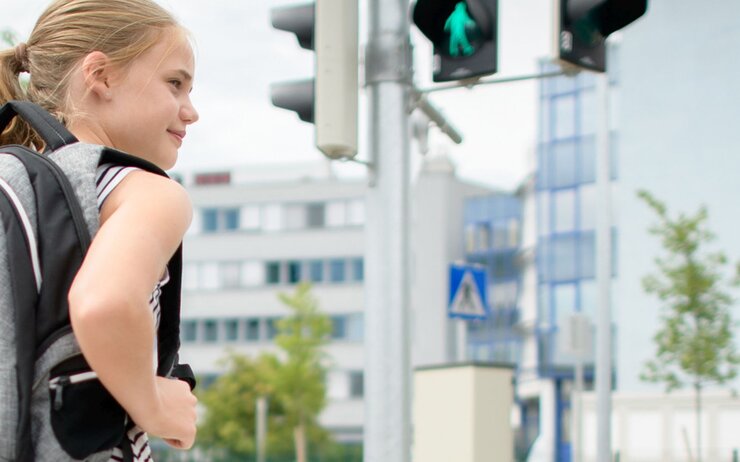 Schulweg üben Ein Mädchen steht an einer Ampel, sie trägt einen schwarz-grauen Rucksack und ist seitlich von hinten zu sehen. Im Hintergrund sind eine Fußgängerampel und eine Kreuzung mit großen Firmengebäuden zu sehen..