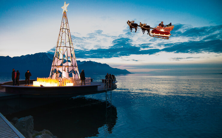 Weihnachtsmarkt Montreux Ein beleuchteter Weihnachtsbaum auf einem Steg am Meer, mit einem fliegenden Weihnachtsmannschlitten im Hintergrund.