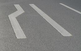 Weiße Bodenmarkierungen auf der Straße, die eine Fahrbahnverengung anzeigen.