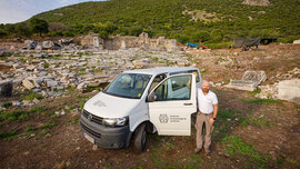 Archäologe Simon Hye steht vor dem weißen VW-Bus, mit dem er nach Ephesos gefahren ist. Im Hintergrund sind Ruinen zu sehen.