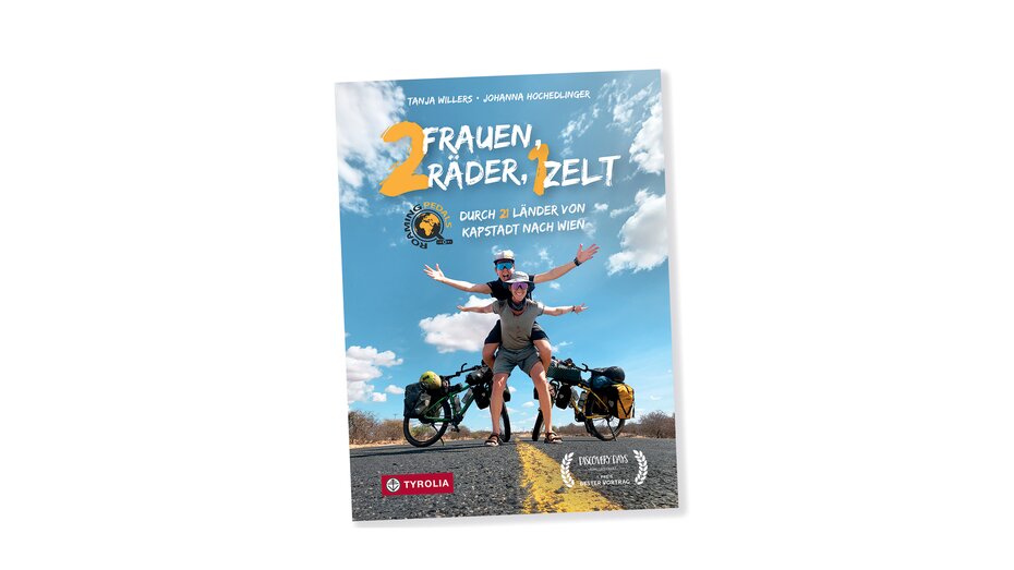 Buchcoverbild "2 Frauen, 2 Räder, 1 Zelt". Darauf sind 2 Frauen zu sehen, die vor ihren Rädern auf einer Straße stehen. Die eine hat die andere huckepack am Rücken.