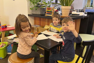 Drei Kids sitzen im Spiele-Café am Tisch. Ein Kind winkt.