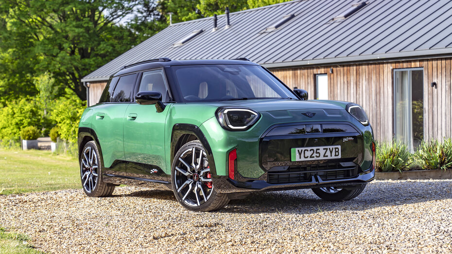 Ein grüner Mini JCW Aceman steht auf einem Schotterplatz vor einem Holzhaus.