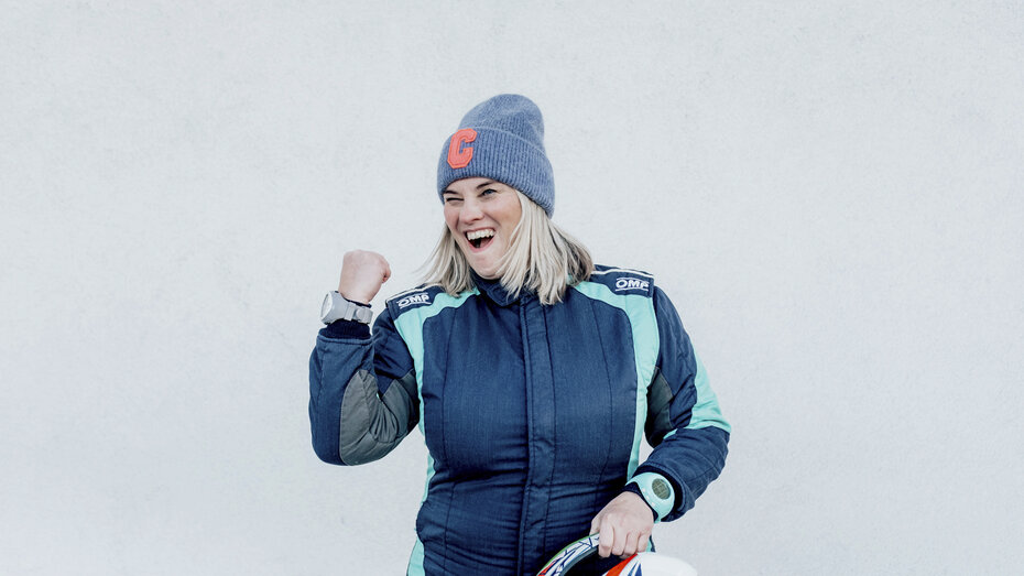 Catharina Schmidt, Rallye-Co-Pilotin und ÖAMTC-Mitarbeitetin steht im Renndress vor hellgrauem Hintergrund in Siegespose und mit Haube.