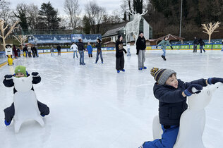 Zwei Kinder sitzen am Eisbären auf dem Eis und haben Eislaufschuhe an.