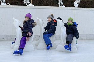 Drei Kinder sitzen am Eisbären auf dem Eis und haben Eislaufschuhe an.