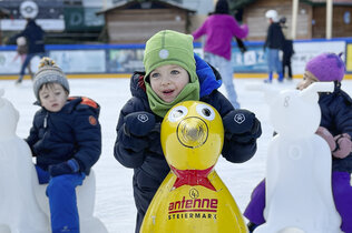 Drei Kinder sitzen am Eisbären auf dem Eis und haben Eislaufschuhe an.