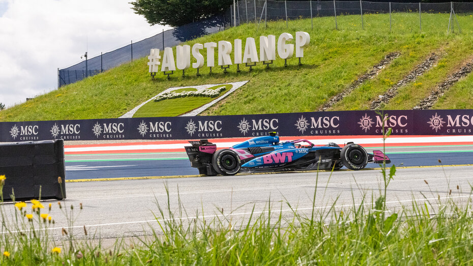 Ein blau-rosa Alpine-F1-Rennauto fährt durch eine Kurve auf einer Rennstrecke, im Hintergrund befindet sich ein "Austrian GP"-Schriftzug auf einer grünen Wiese.