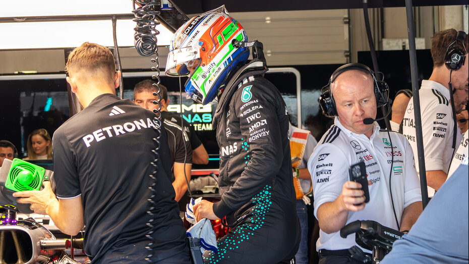 Ein Formel-1-Fahrer in voller Montur mit Rennanzug und -helm steht neben seinem Formel 1 Auto, um ihn herum arbeiten viele Teammitglieder.