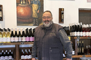 Ein Weinbauer mit grauem Gilet lacht in die Kamera, im Hintergrund Weinflaschen im Regal.