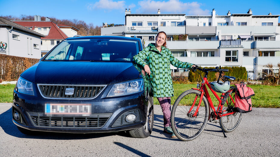 Eine Frau steht in der MItte zwischen einem schwarzen Auto und einem Fahrrrad. Im Hintergrund ein Wohngebäude.