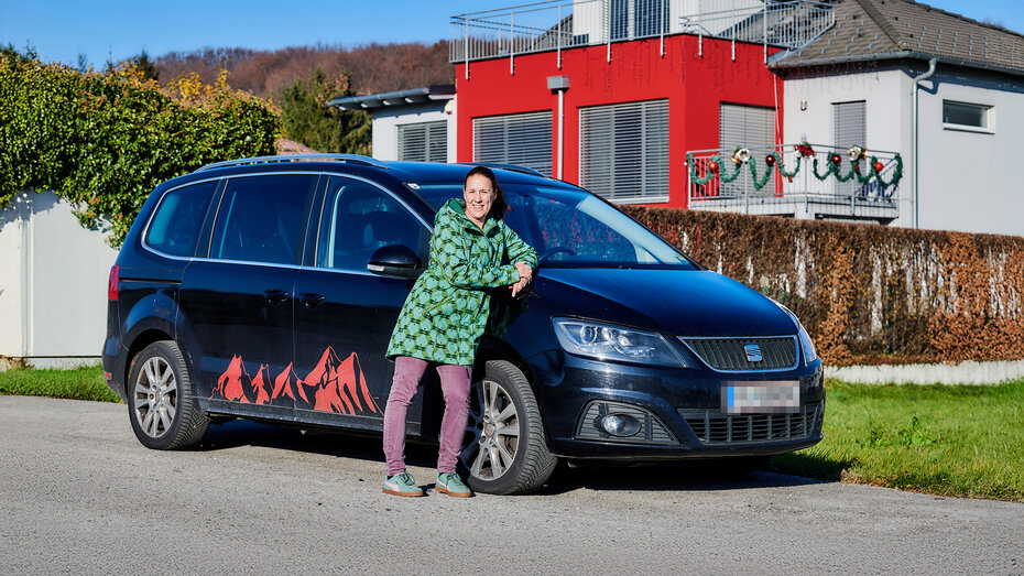 Eine Frau, die Hebamme Martina Klasz, steht an ihr schwarzes Auto gelehnt und lächelt. Im Hintergrund ein Wohnhaus.