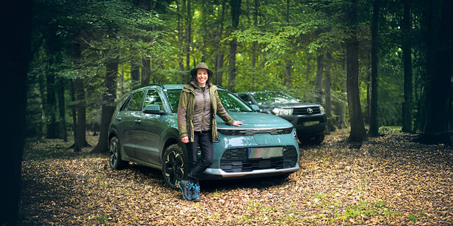 Eine Försterin lehnt an ihrem Auto, das mitten im Wald steht. Dahinter steht ein weiteres Fahrzeug.