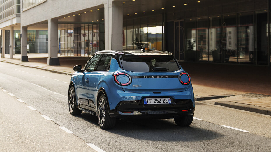 Ein blauer Nissan Micra 2025 fährt auf einer Straße an einem modernen Gebäude vorbei.