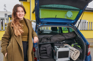 Sängerin Anna Pflamitzer posiert vor ihrem blauen Auto, das vor einem gelben Haus geparkt ist. Der Kofferraum ist geöffnet und zeigt ihr Arbeitsmaterial.