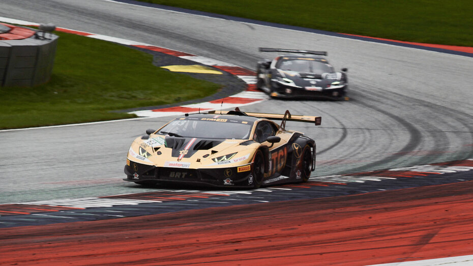 Ein gold-schwarzer Lamborghini-Rennwagen fährt über die Curbs nach einer Kurve auf einer Rennstrecke, dahinter fährt ein dunkelblaues Rennauto.