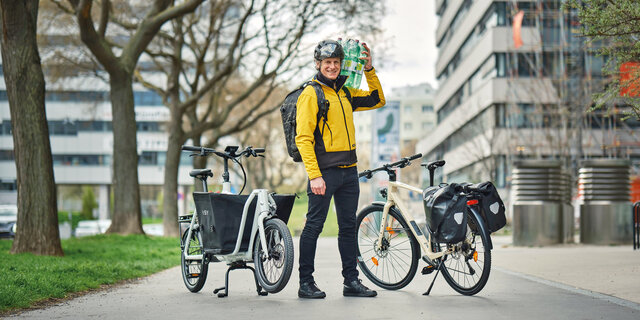 Redakteur mit gelber Jacke und geschulterten Wasserflaschen steht zwischen einem Lastrad und einem Fahrrad mit Gepäcktaschen.