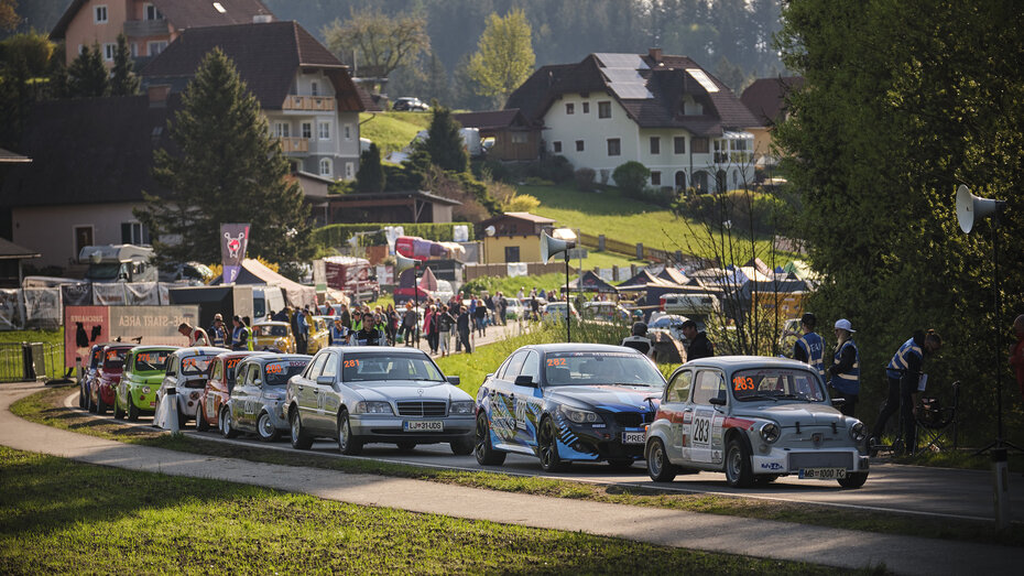 Rennautos stehen auf einer Landstraße, drumherum gehen Menschen.