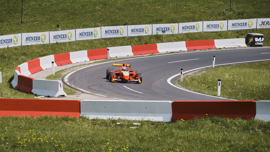 Ein rot-weißes-Formel-Rennauto fährt durch eine Kurve auf einer Landstraße.