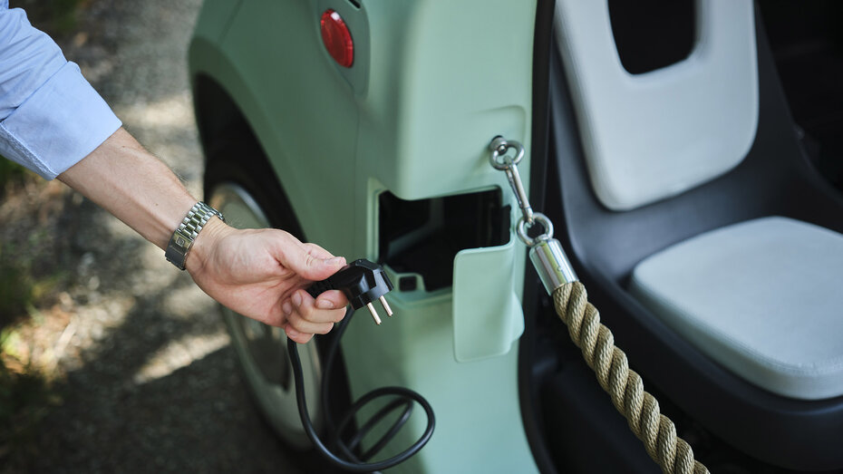 Eine Hand hält ein Ladekabel in der Hand, das in der Tür eines türkisen Microcars integriert ist.