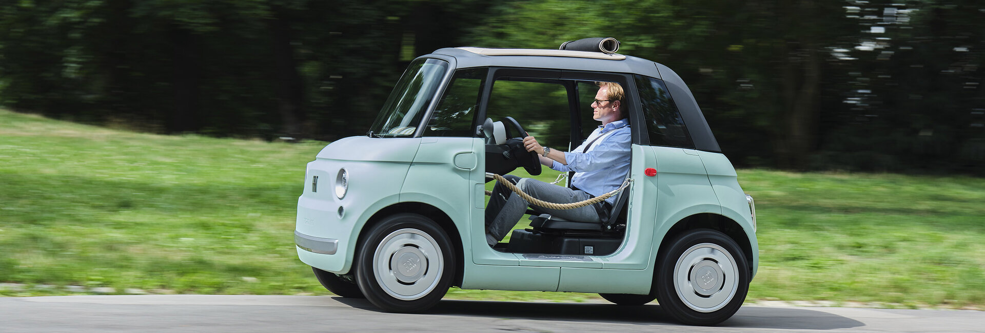 Eine Person fährt mit einem türkis-schwarzen Microcar, das anstelle von Türen braune Flechtkordeln hat, über eine Straße, die an einer grünen Wiese und Bäumen entlang führt.