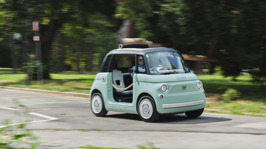 Eine Person fährt mit einem türkis-schwarzen Microcar, das anstelle von Türen braune Flechtkordeln hat, über eine Straße, die an einer grünen Wiese und Bäumen entlang führt.
