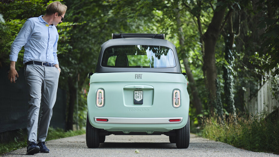 Eine Person mit grauer Hose und blauem Hemd entfernt sich von einem türkisen Microcar steht auf einer Straße, die von Bäumen umgeben ist, und blickt auf das Fahrzeug zurück.