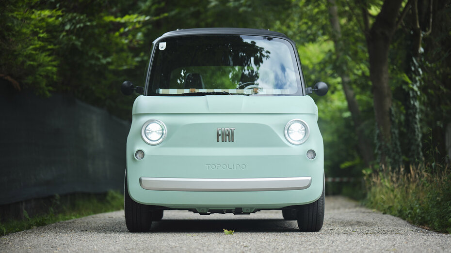 Ein türkises Microcar steht auf einer einspurigen Straße, die von Bäumen umgeben ist.