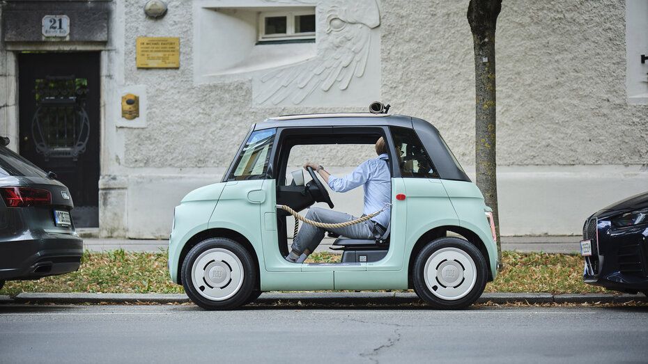Eine Person parkt ein türkises Microcar, das anstelle von Türen Flechtkordeln hat, längs ein.