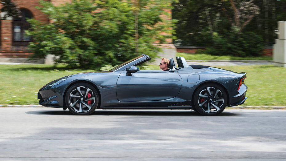 Ein graues Cabrio fährt mit offenem Verdeck auf einer Straße an einer Wiese vorbei.