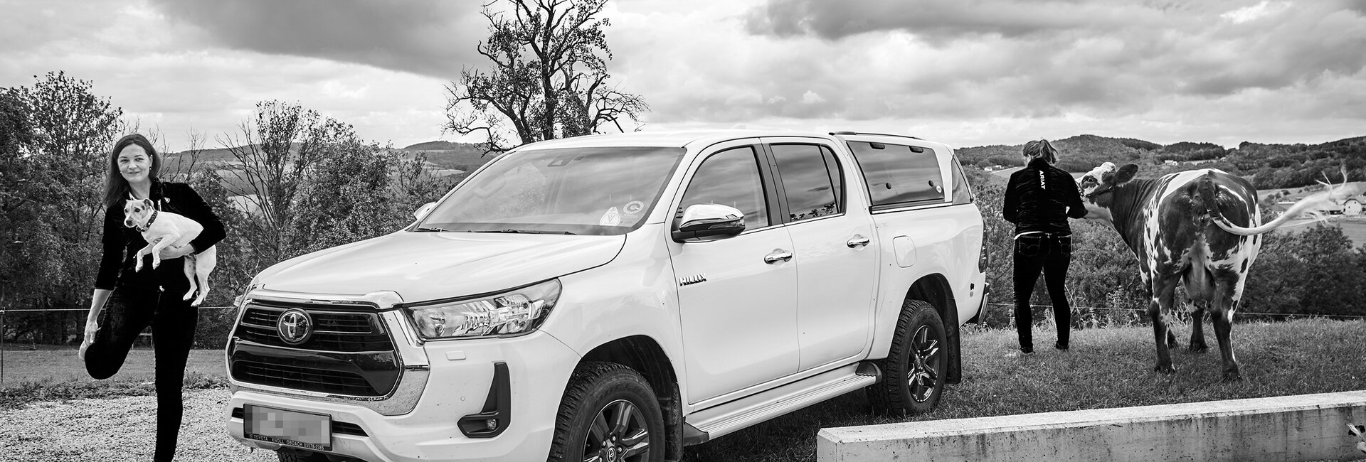 Die Tierärztinnen Natalie und Stephanie Debeerst (mit einer Kuh an der Leine) und ihr Auto. Natalie schaut ins Bild und hat einen Hund in der Hand.