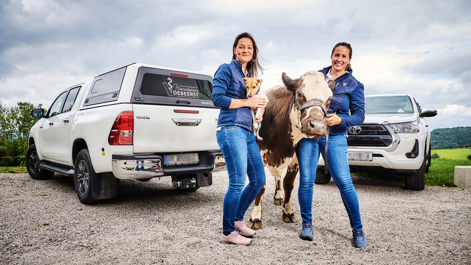 Zwei Frauen stehen mit einer Kuh, und einem Hund vor ihren Pick-ups.