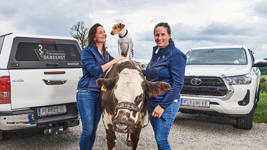 Die Tierärztinnen posieren mit ihrer Kuh und ihrem Hund vor zwei Pick-ups. Der Hund sitzt auf der Kuh, die Tierärztin schaut ihn an.