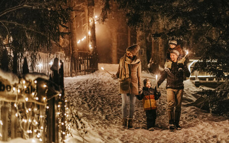 Eltern gehen mit zwei Kindern im Schnee im Wald. Die Werg ist mit beleuchtet.
