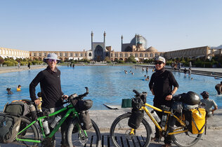 Zwei Frauen mit Hut und Sonnenbrille stehen mit ihrem bepackten Rädern vor dem Wasserbassin der Moschee in Isfahan, die im Hintergrund zu sehen ist.