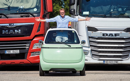 Eine Person steht aufrecht und mit ausgestreckten Armen im hellgrünen Kleinfahrzeug Microlino und blickt durch das Schiebedach. Dahinter stehen ein roter und weißer Lkw.