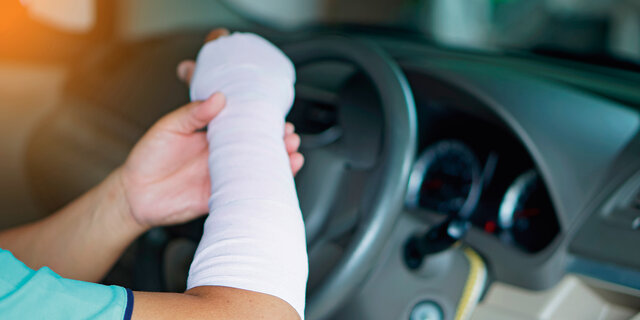 Ein verletzter Mann mit einem Verband an Hand und Arm fährt ein Auto, was zu einem Unfall führen könnte, weil es unbequem ist, wie mit einer normalen Hand zu fahren.