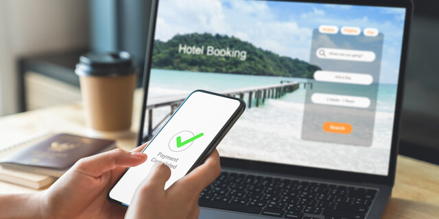 Hand vor einem Laptop mit offener Website zur Hotelbuchung für den Urlaub. Handy zeigt als Nachricht "abgeschlossene Zahlung" an.