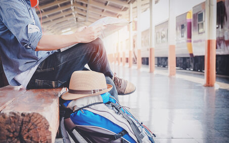 Ein Bahnreisender sitzt mit einer Zeitung und einem Rucksack am Bahnsteig und wartet auf den Zug.