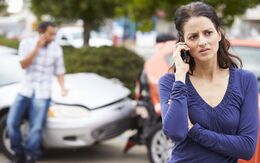Im Vordergrund sieht man eine junge Frau mit besorgtem Gesichtsausdruck, die per Handy telefoniert. Im Hintergrund einen Mann vor zwei Autos, die einen Auffahrunfall verursacht haben.
