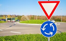 Kreuzung mit Kreisverkehr an einem sonnigen Tag. Im Vordergrund ist ein Vorrang geben-Verkehrsschild zu sehen mit einer blauen, runden Zusatztafel, die den Kreisverkehr anzeigt.